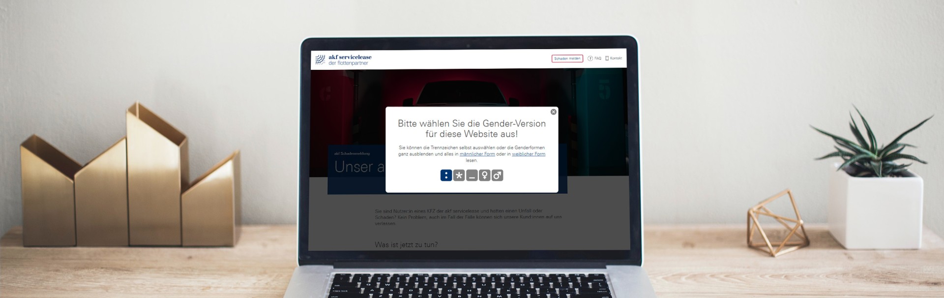 Ein aufgeklappter, silberner Laptop steht auf einem Holztisch. Auf dem Bildschirm ist die Startseite der Schadenmeldung-Website zu sehen, mit dem weißen Abfragefenster des Gender Selector im Vordergrund. Neben dem Laptop stehen Deko-Gegenstände aus Metall und eine Topfpflanze.