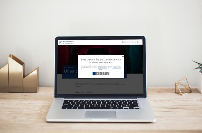 Ein aufgeklappter, silberner Laptop steht auf einem Holztisch. Auf dem Bildschirm ist die Startseite der Schadenmeldung-Website zu sehen, mit dem weißen Abfragefenster des Gender Selector im Vordergrund. Neben dem Laptop stehen Deko-Gegenstände aus Metall und eine Topfpflanze.