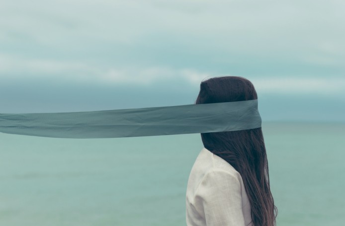 Eine Frau in hellem Oberteil mit langen, braunen Haaren ist von der Seite zu sehen. Ein schmales, blaues Tuch bedeckt ihre Augen und wird waagrecht nach hinten weggeweht. Im Hintergrund ist verschwommen eine Wasseroberfläche und bewölkter Himmel zu sehen.