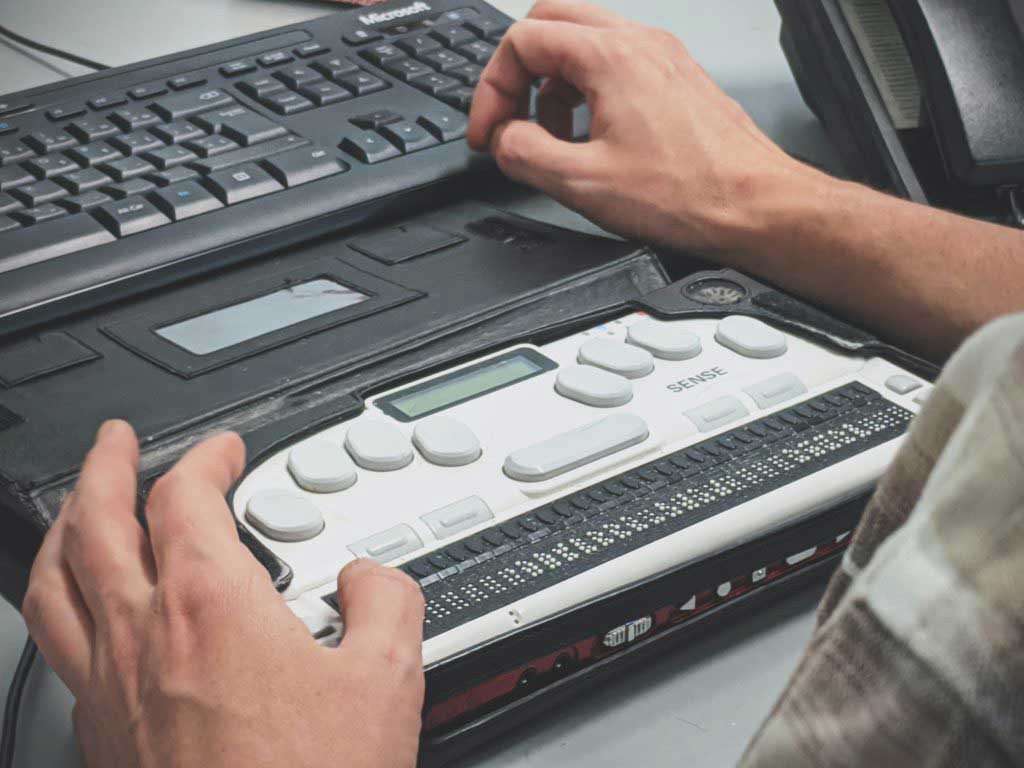 Eine Braille-Zeile steht vor einer Computertastatur, zwei Hände bedienen sie.
