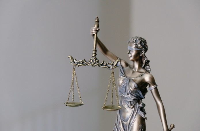 Eine Metallstatue der römischen Göttin Justitia vor hellem Hintergrund. Mit dem rechten Arm hält sie eine Waage hoch. Die beiden Waagschalen befinden sich auf gleicher Höhe. Die Augen der Göttin sind mit einem Tuch verbunden.