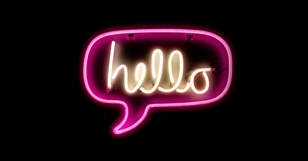 Vor einem schwarzen Hintergrund leuchtet eine pinkfarbene Sprechblase aus Neonröhren. Darin steht in weißer Schrift das Wort hello.