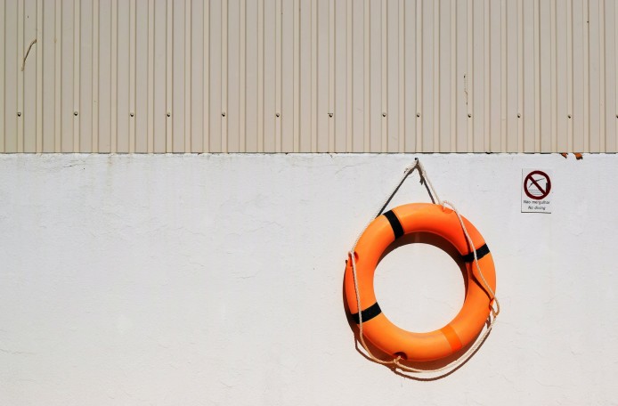 Ein orangefarbener Rettungsring hängt an einer hellgrauen Wand. Daneben hängt ein Verbotsschild mit der Aufschrift Nao mergulhar - No diving.